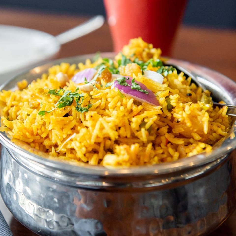 Close-up of Tandoori Flame's yellow biryani rice, garnished with red onions and cilantro in a metal bowl.