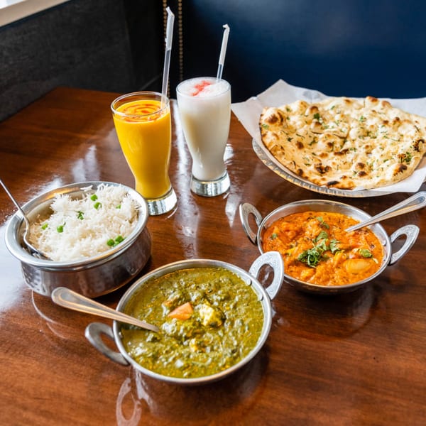 Indian meal with naan, rice, two curries, mango lassi, and sweet lassi on a wooden table.